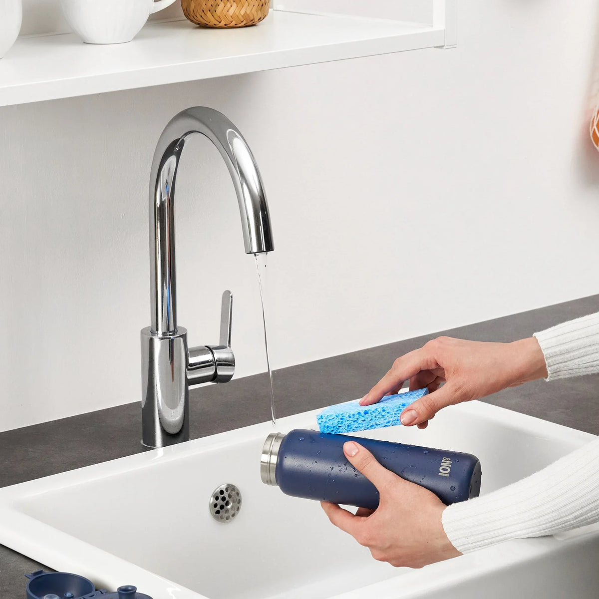Hands scrubbing a navy bottle with a blue sponge under a running faucet in a kitchen sink context.