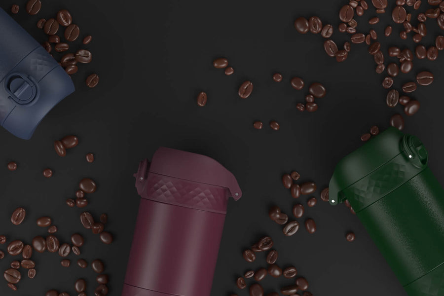 Several portable coffee makers, in various colors, are scattered on a dark surface amongst coffee beans.