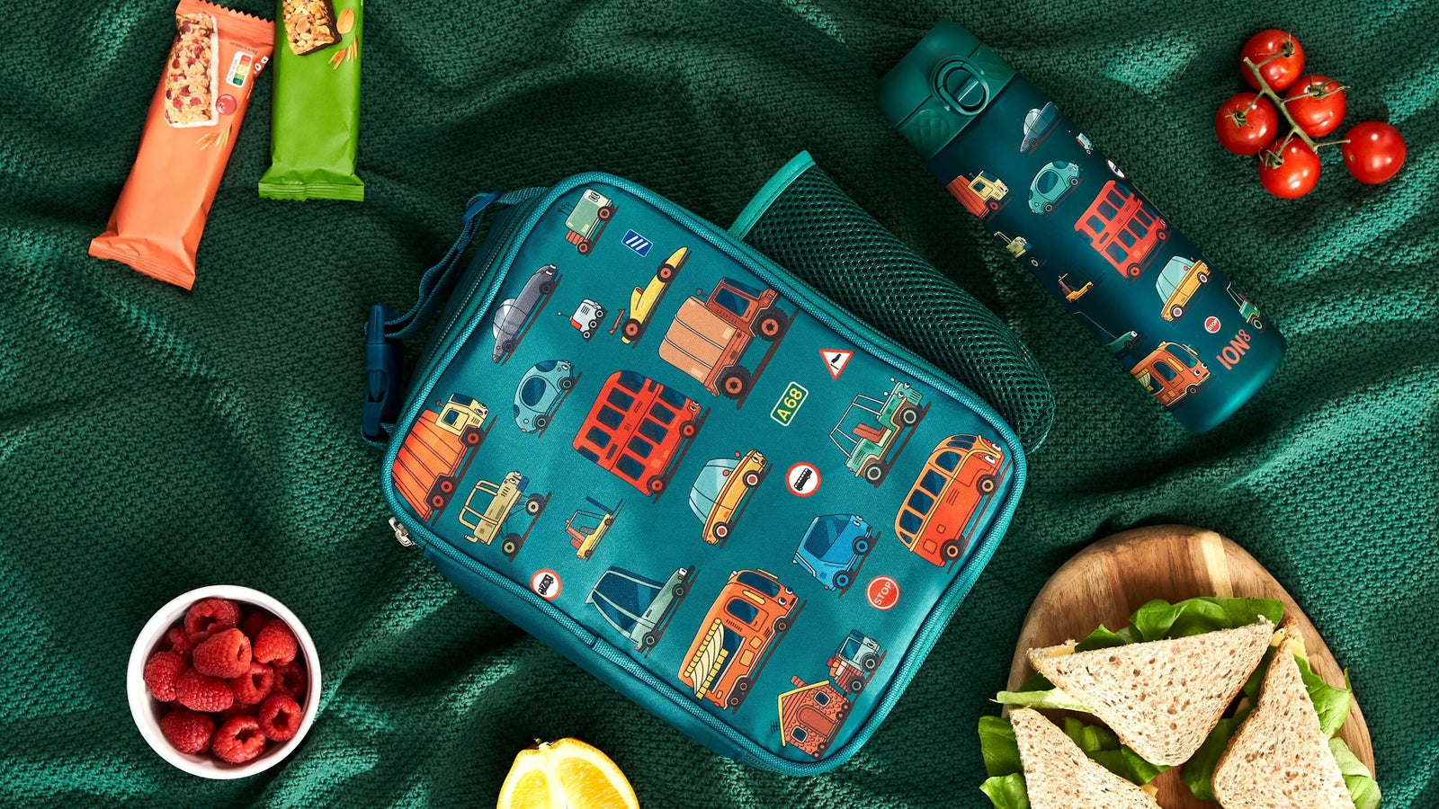 A teal lunch bag, adorned with vehicle illustrations, rests on a green fabric. Nearby are food items and a matching water bottle labeled "ION8".