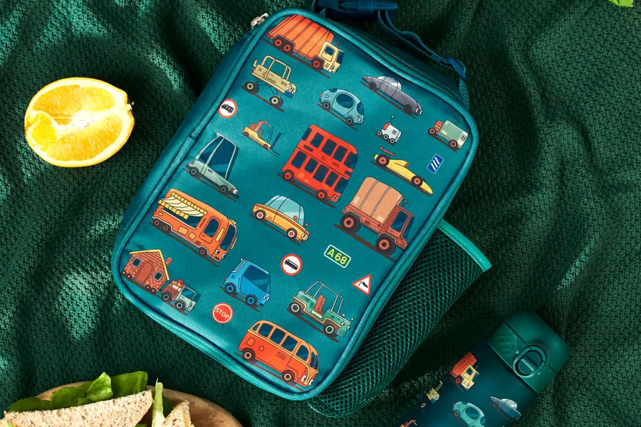 A teal lunch bag, decorated with various vehicles, rests on a green blanket alongside a matching water bottle, sandwiches, fruit, and granola bars.